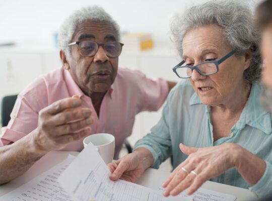 Älteres Paar bespricht Finanzunterlagen mit einem Berater als Symbol für Pensionsverpflichtungen, Altersvorsorge und langfristige Unternehmenszusagen.