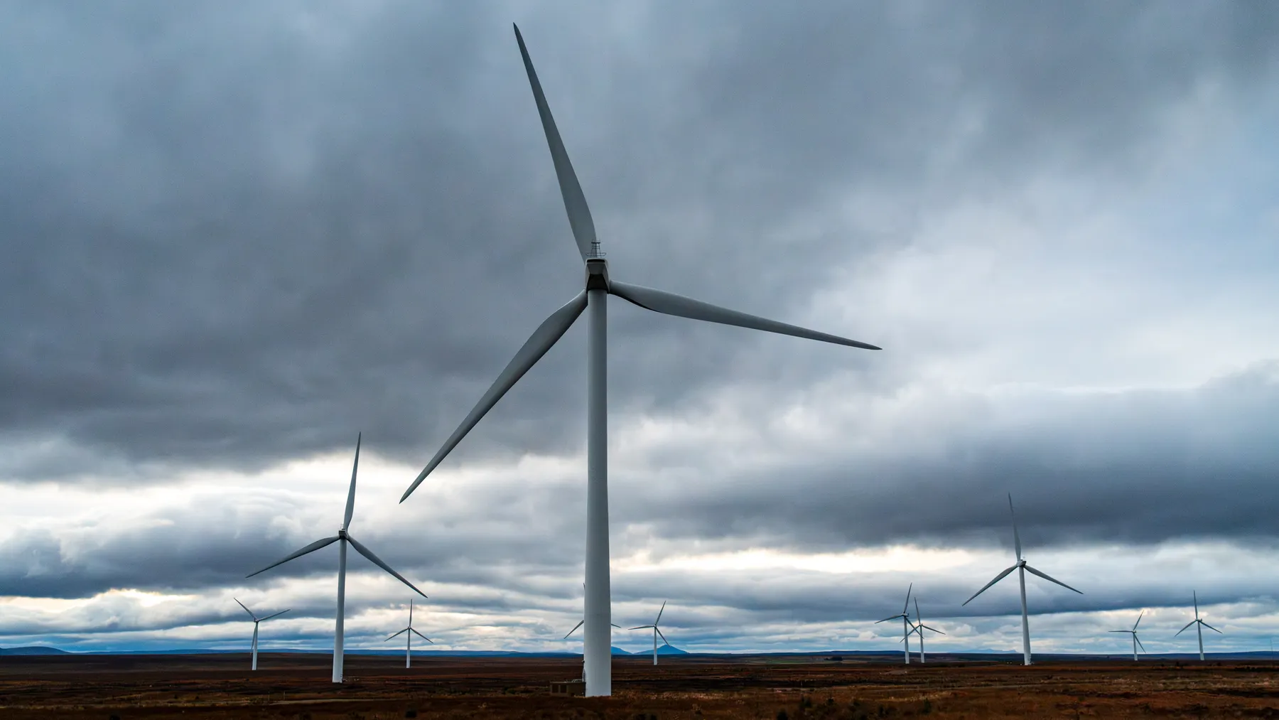 Mehrere moderne Windenergieanlagen auf weitläufigen Feldern unter dramatischen Wolken; Symbolfoto für Risiken im Windpark-Projektgeschäft.