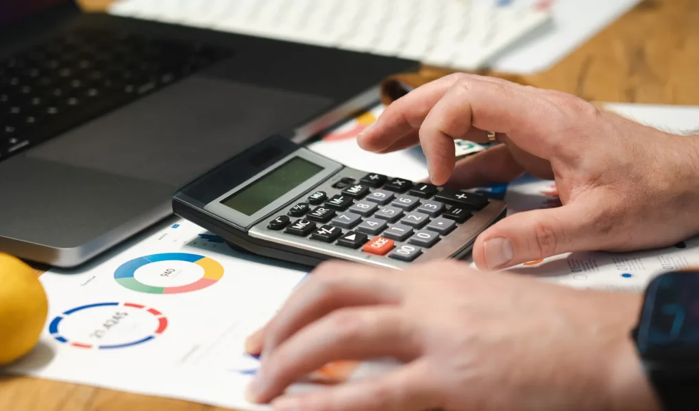 Hände prüfen Finanzunterlagen mit Taschenrechner und Laptop auf einem Schreibtisch.