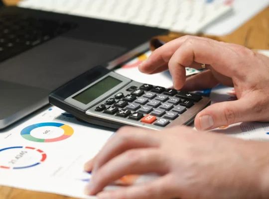 Hände prüfen Finanzunterlagen mit Taschenrechner und Laptop auf einem Schreibtisch.