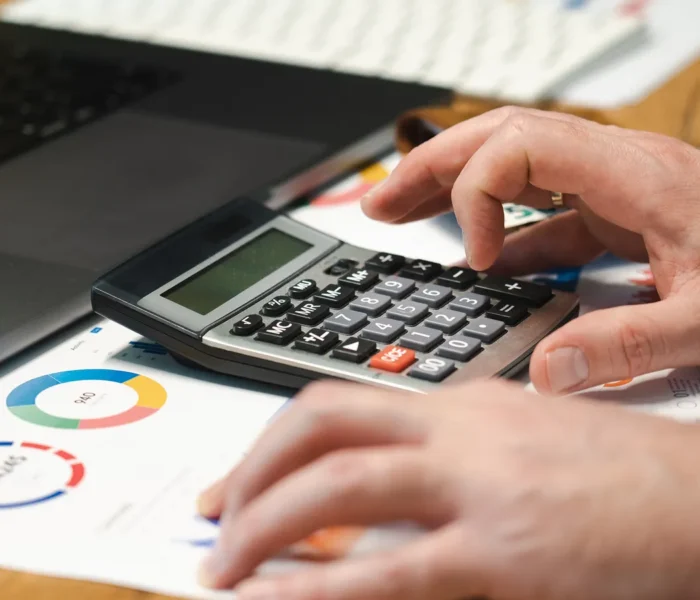 Hände prüfen Finanzunterlagen mit Taschenrechner und Laptop auf einem Schreibtisch.