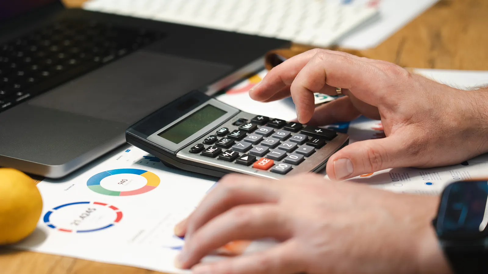 Hände prüfen Finanzunterlagen mit Taschenrechner und Laptop auf einem Schreibtisch.